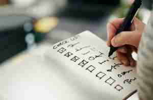 A hand holds a pen and checks off items on a handwritten checklist in a notebook. The items have checkboxes, and several have already been marked as completed. The writing is mostly in black ink.