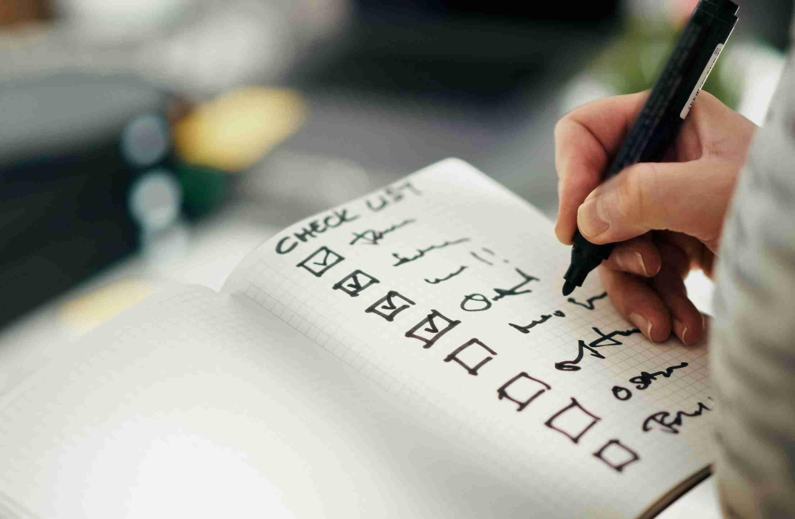 A hand holds a pen and checks off items on a handwritten checklist in a notebook. The items have checkboxes, and several have already been marked as completed. The writing is mostly in black ink.