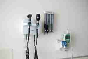 Medical instruments mounted on a white wall, including an otoscope, ophthalmoscope, blood pressure cuff, and a thermometer holder, typically found in a doctor's examination room.