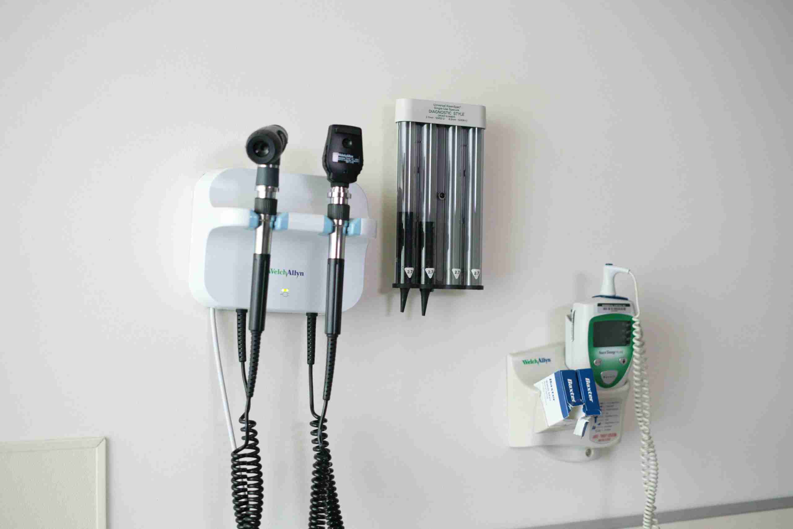 Medical instruments mounted on a white wall, including an otoscope, ophthalmoscope, blood pressure cuff, and a thermometer holder, typically found in a doctor's examination room.