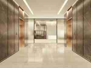 A modern elevator lobby with four closed elevators, marble floors, and metallic wall panels; in the background, glass doors reveal a minimalistic reception area with a marble desk.