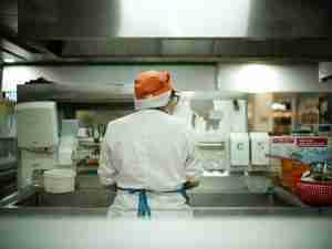 A person wearing a white chef’s coat, orange hat, and blue apron stands at a kitchen sink, facing away from the camera. The kitchen area contains various equipment and supplies.