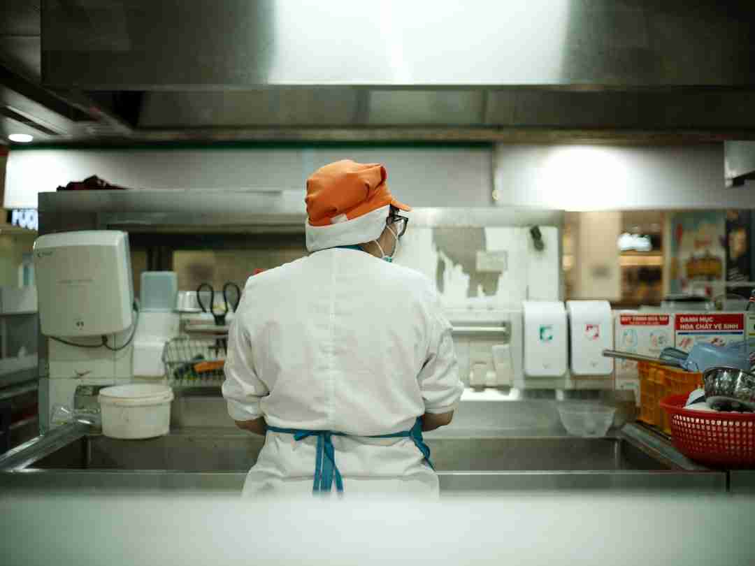A person wearing a white chef’s coat, orange hat, and blue apron stands at a kitchen sink, facing away from the camera. The kitchen area contains various equipment and supplies.