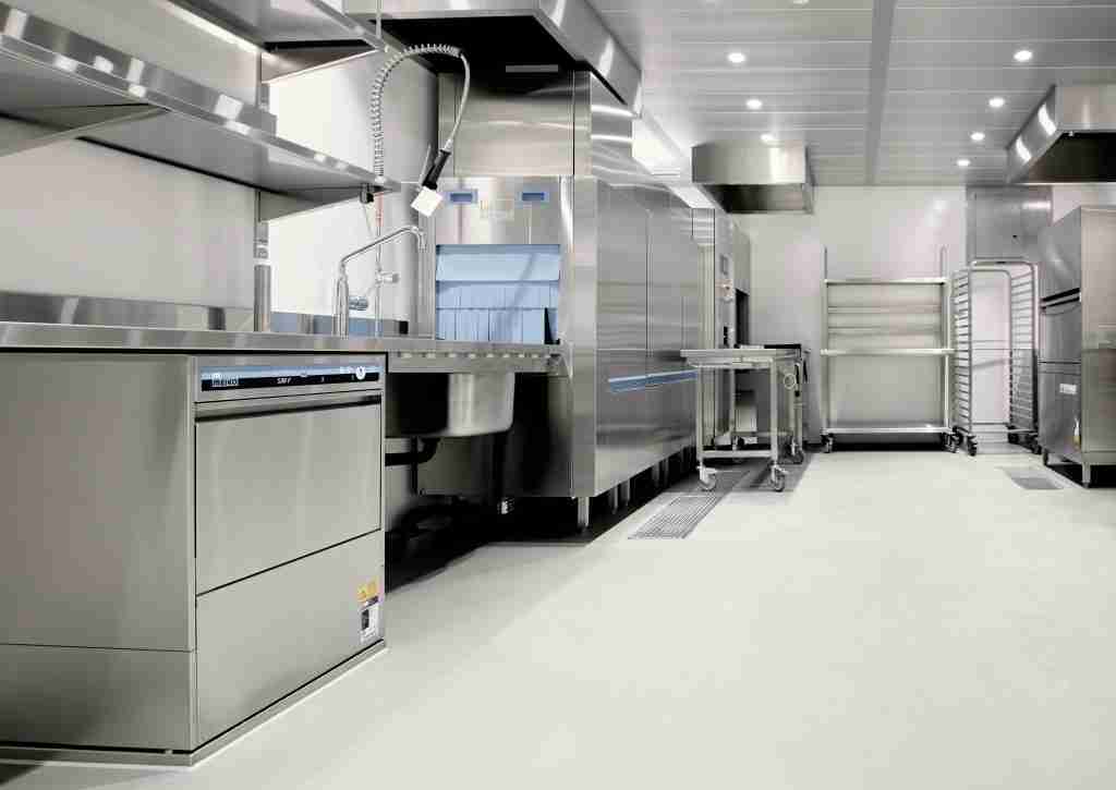 A clean, modern commercial kitchen featuring stainless steel appliances, a dishwasher, industrial sinks, metal tables, drying racks, and bright overhead lighting. The area appears empty and well-organized.