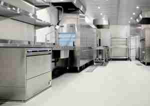 A clean, modern commercial kitchen featuring stainless steel appliances, a dishwasher, industrial sinks, metal tables, drying racks, and bright overhead lighting. The area appears empty and well-organized.