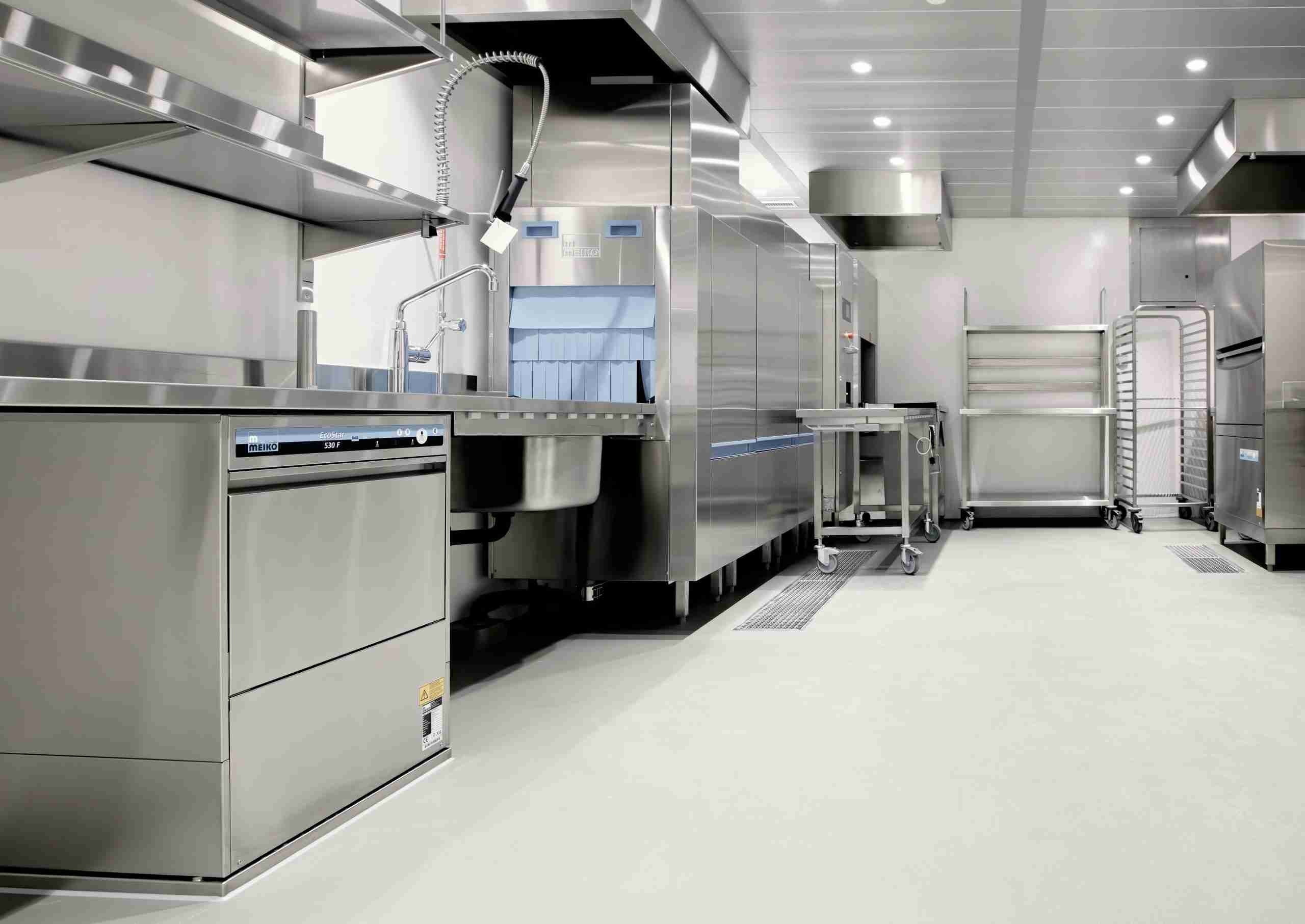 A clean, modern commercial kitchen featuring stainless steel appliances, a dishwasher, industrial sinks, metal tables, drying racks, and bright overhead lighting. The area appears empty and well-organized.