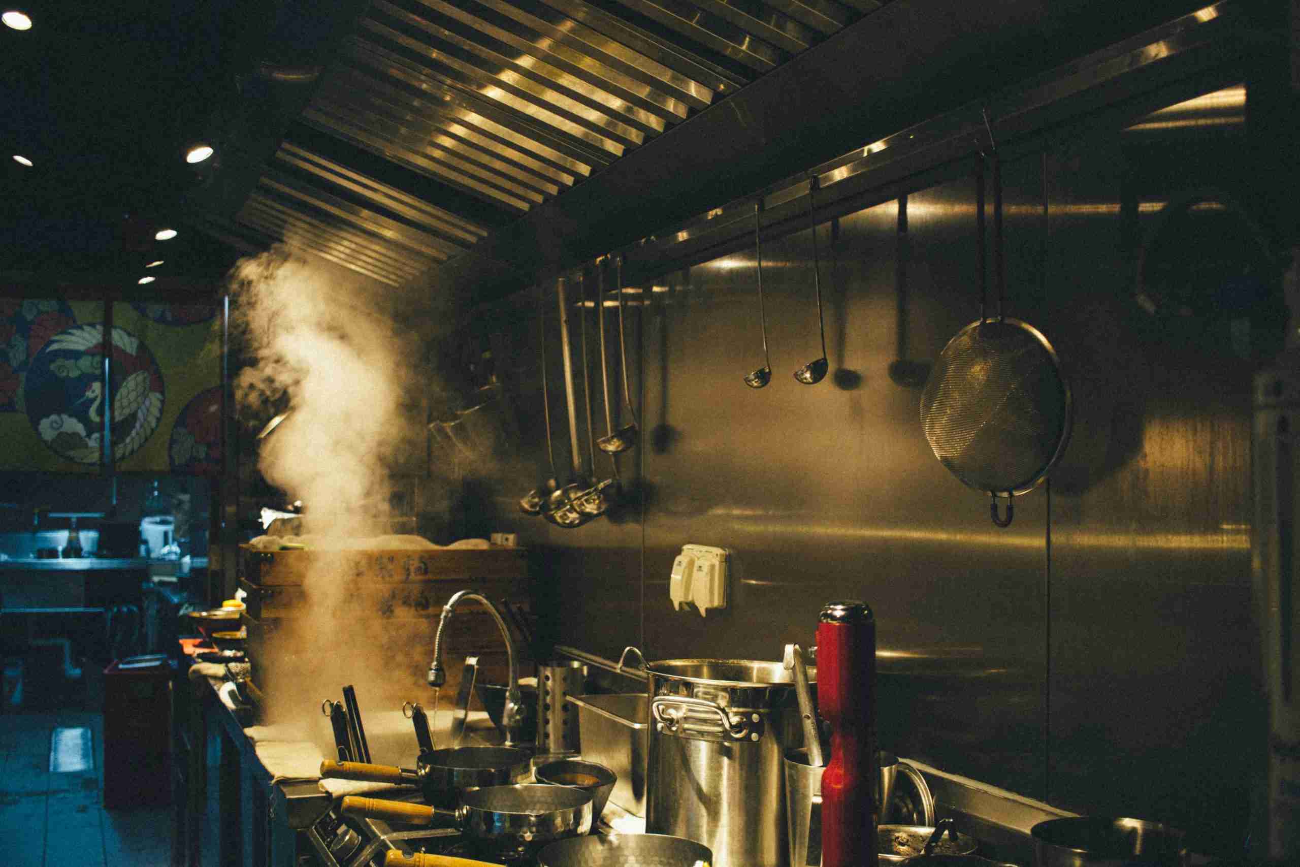 A commercial kitchen with pots on the stove, steam rising, ladles and a pan hanging on a metal wall, and various kitchen tools and equipment visible under warm lighting.