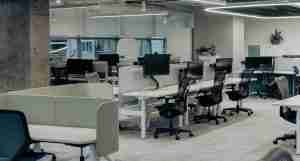 A modern open-plan office with several empty workstations, each equipped with a black office chair, computer monitor, and partition dividers. Large windows provide natural light in the background.