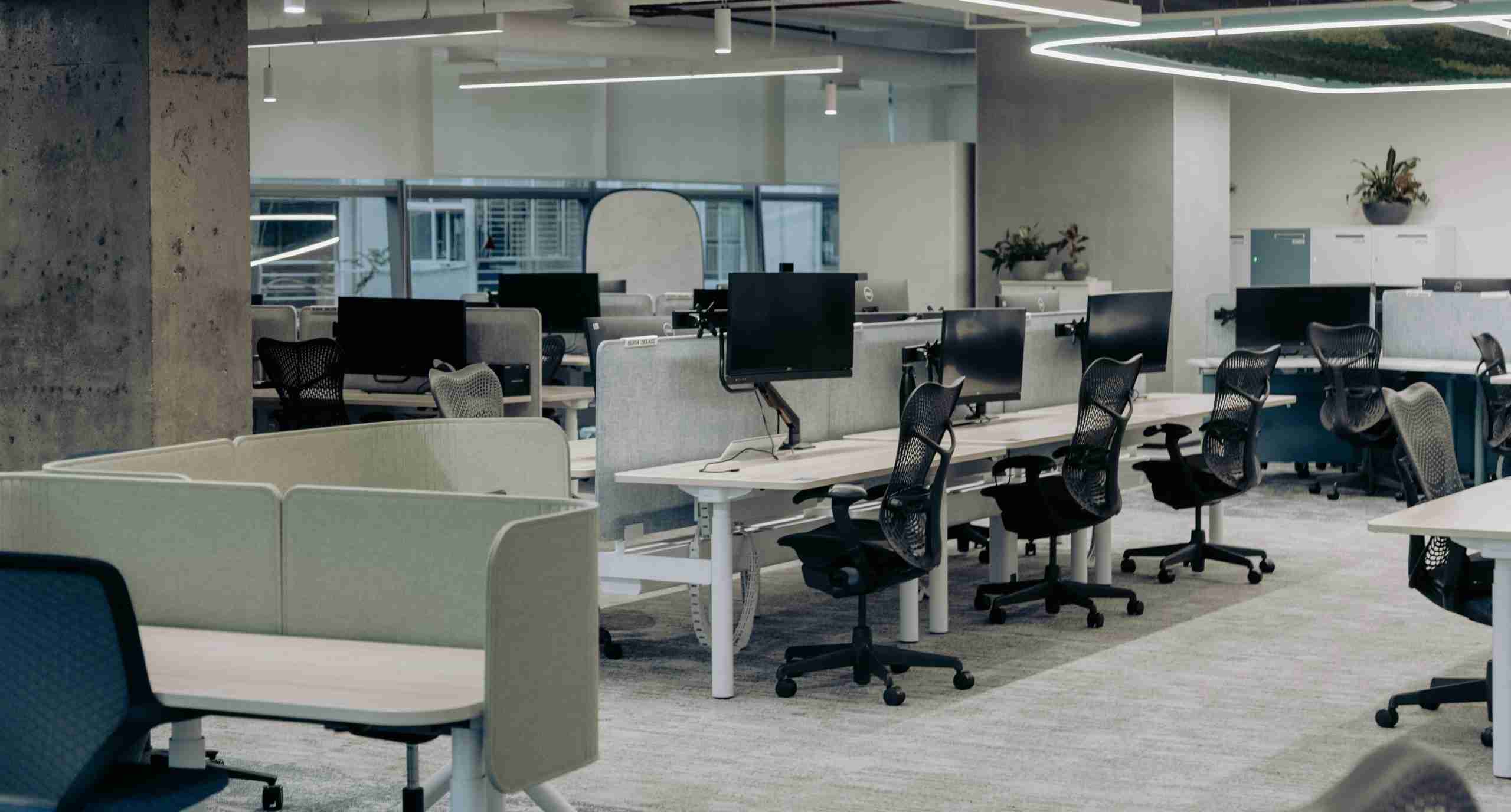 A modern open-plan office with several empty workstations, each equipped with a black office chair, computer monitor, and partition dividers. Large windows provide natural light in the background.