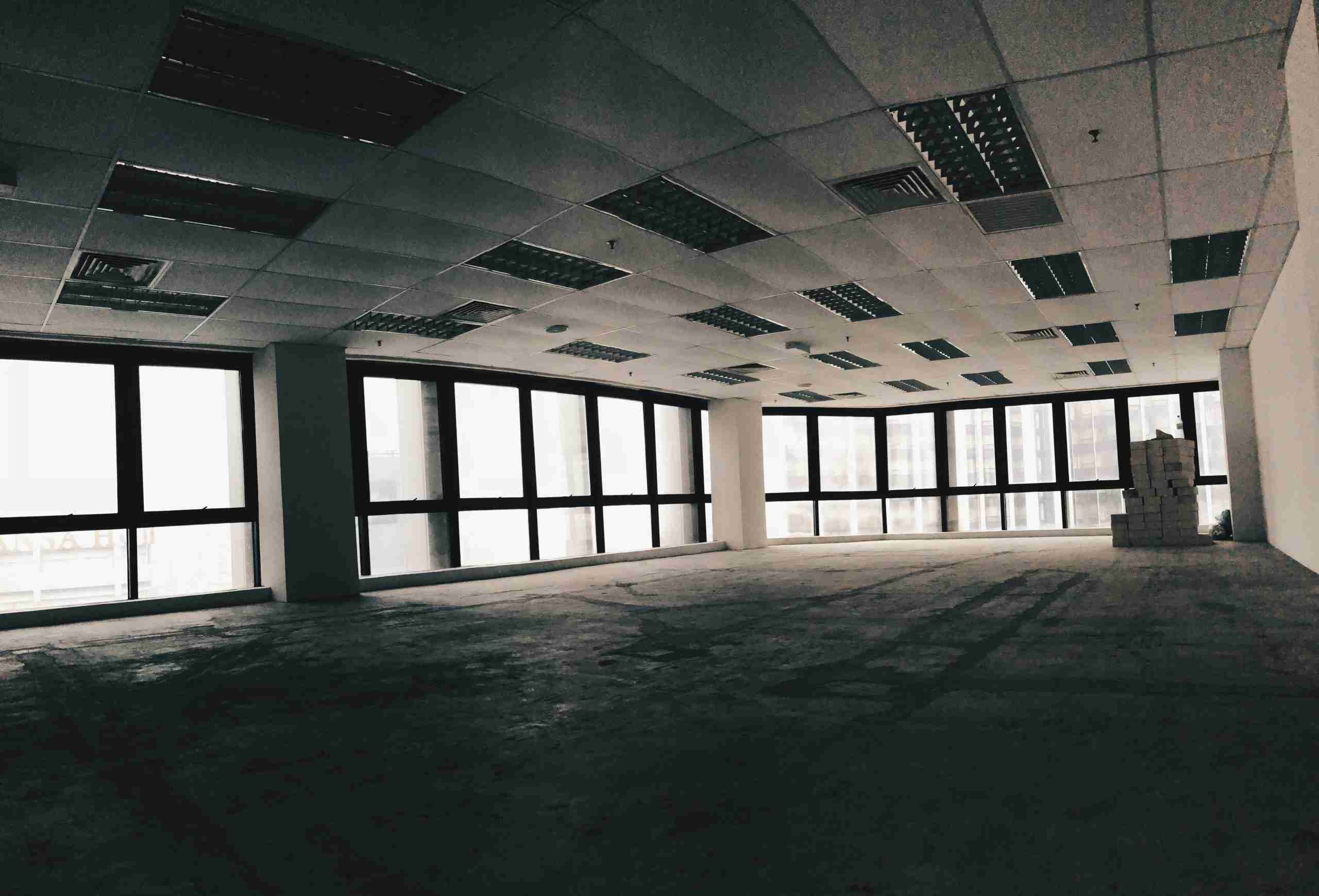An empty office space with large windows along two walls, tiled ceiling panels, fluorescent lights, and an unfinished concrete floor. Stacks of boxes are placed near one corner of the room.