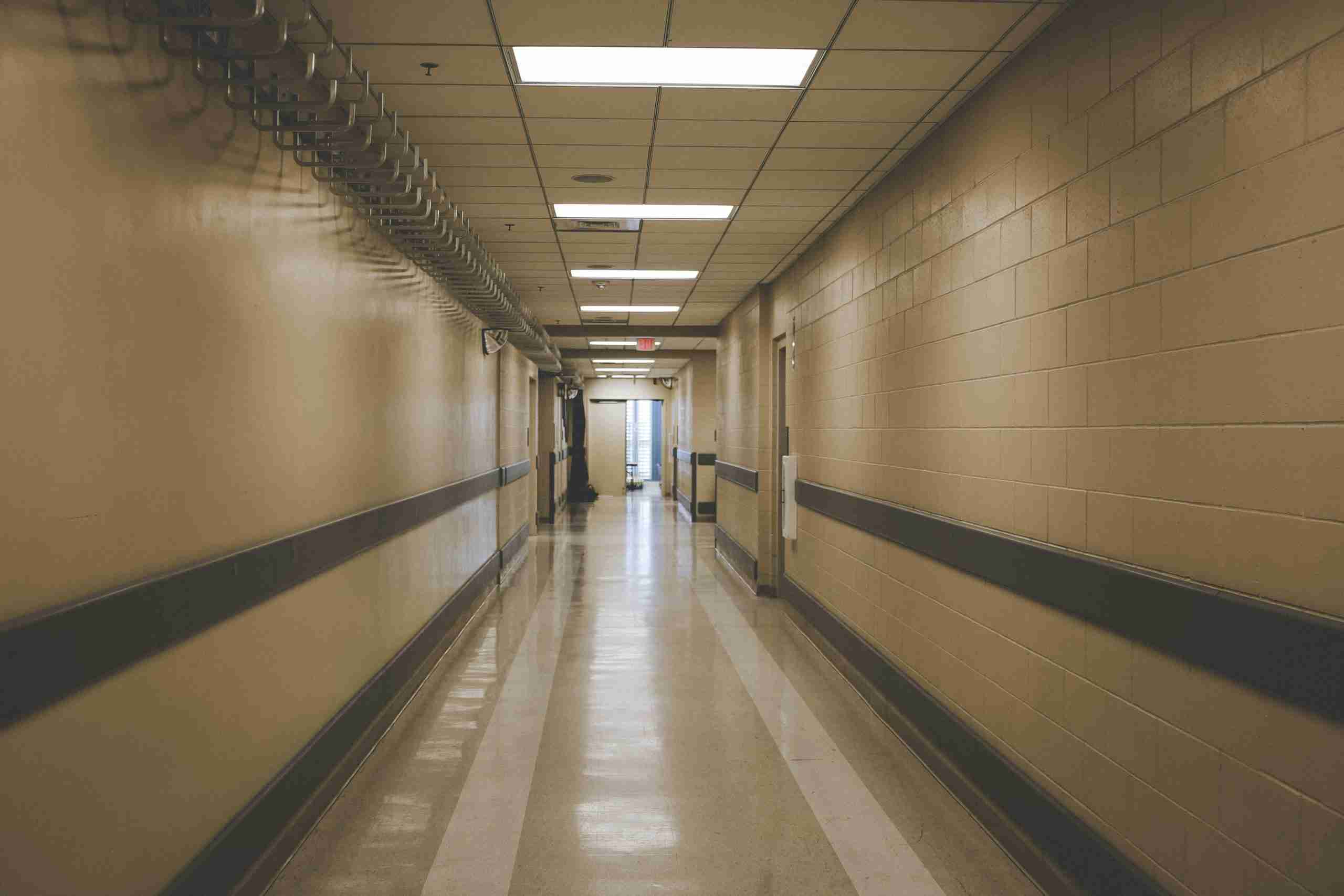 A long, empty hallway with beige walls, fluorescent ceiling lights, and a shiny floor. There are handrails on both sides and pipes along the upper left wall. Double doors are visible at the far end of the hall.