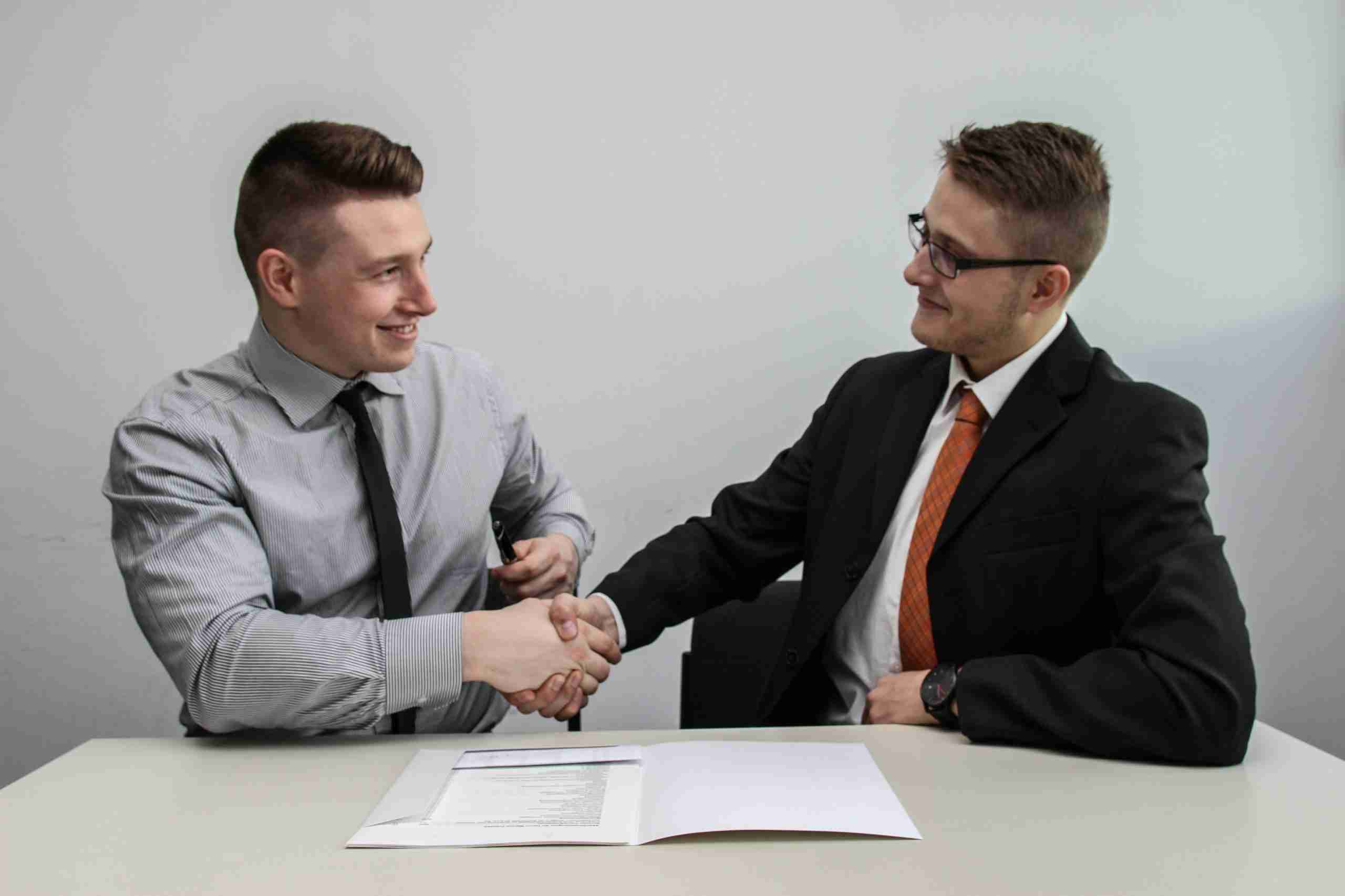 Two men in business attire are sitting at a table, shaking hands and smiling. There are documents on the table in front of them. The background is plain and light-colored.