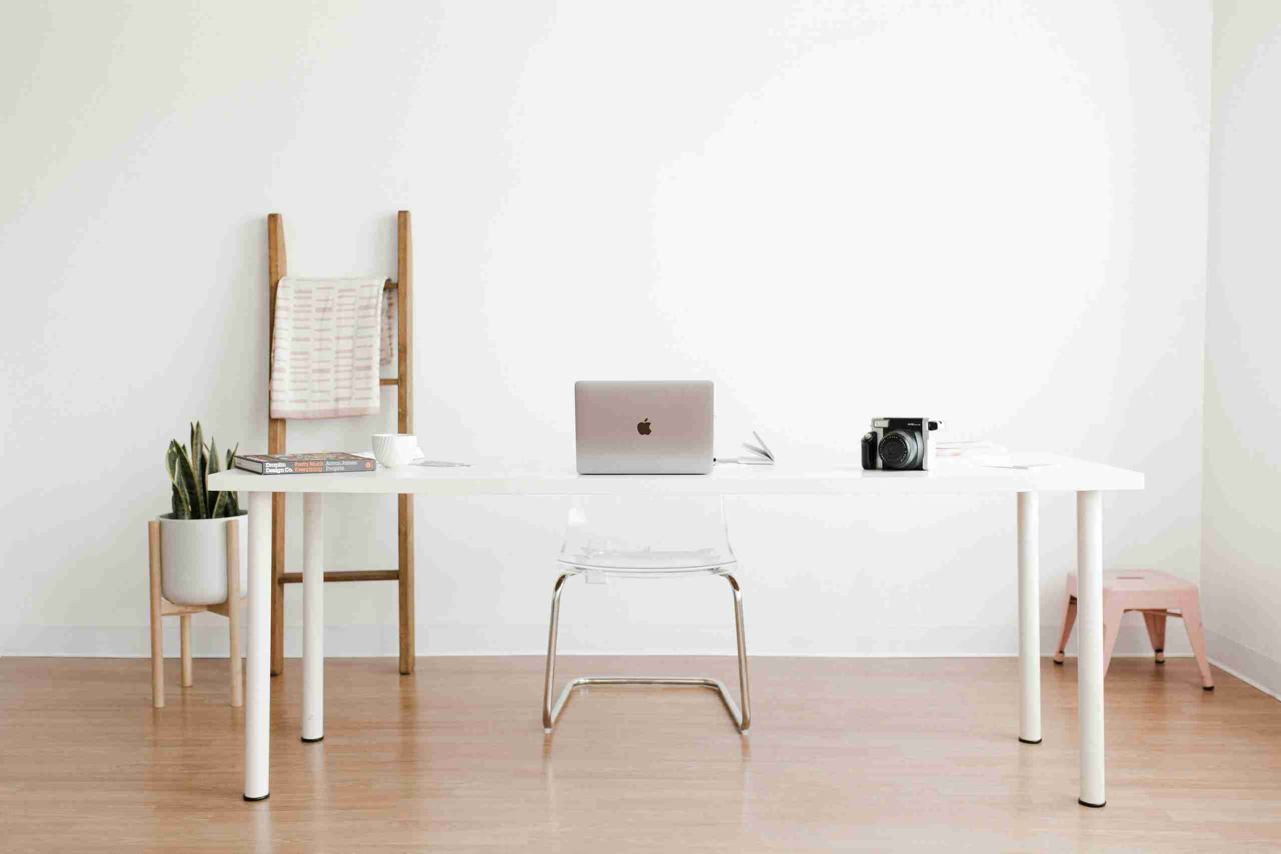 A minimalist office with a white desk, a laptop, a camera, books, and a cup. A clear chair is at the desk. In the background are a plant, a wooden ladder with a draped cloth, and a small pink stool on a light wood floor.