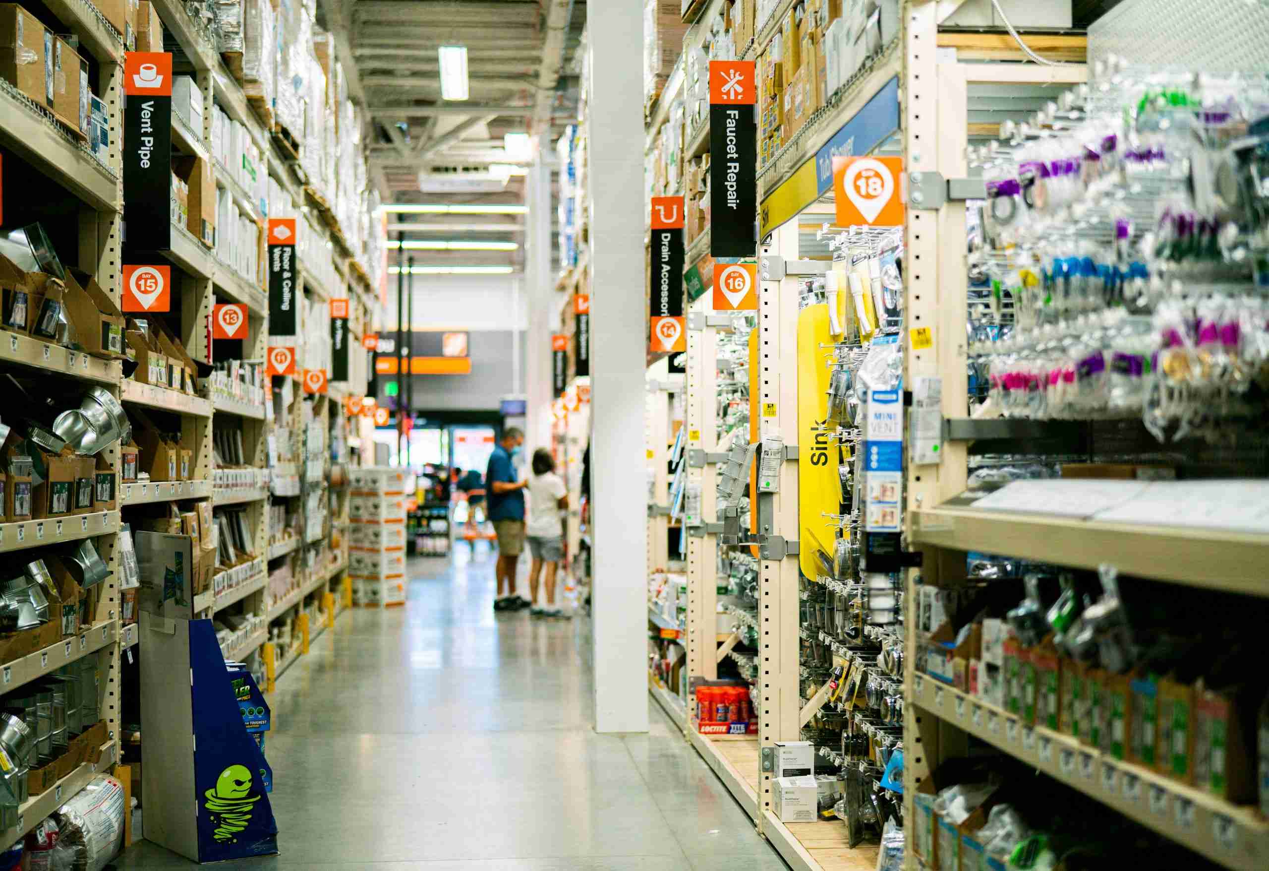 Wide aisle inside a hardware store, lined with shelves stocked with various tools, parts, and plumbing supplies; overhead signs indicate different product categories, and a few people are visible in the distance.