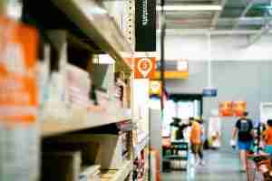 A store aisle with shelves holding various products, a sign marking aisle 3, and several people in the background near the entrance of a large hardware or home improvement store.